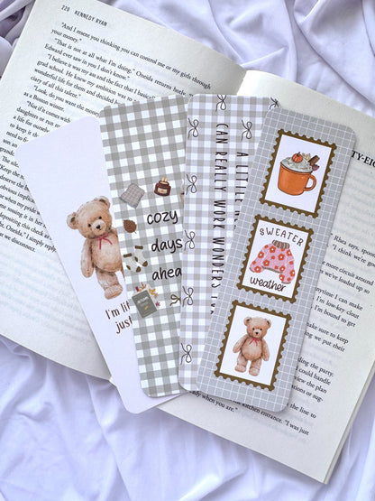 Flat lay of four cozy autumn-themed bookmarks featuring teddy bears, pumpkins, sweater weather illustrations, neutral gingham patterns, and reading quotes displayed on an open book with white bedding in the background.