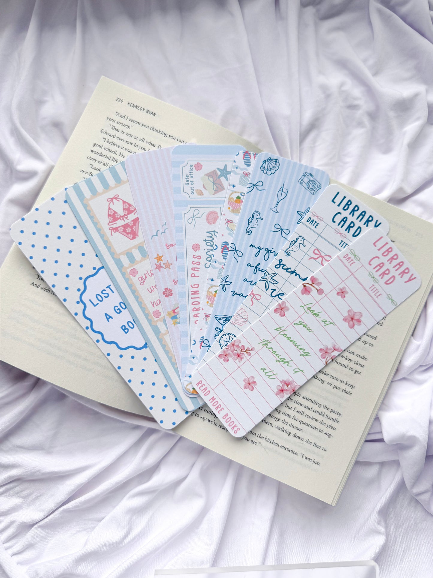 Flat lay of seven summer-themed bookmarks displayed on an open book with white bedding. The bookmark designs feature coastal illustrations, beach icons, pastel colors, library card designs, floral accents, and bookish quotes, creating the perfect reading accessories for summer book lovers.