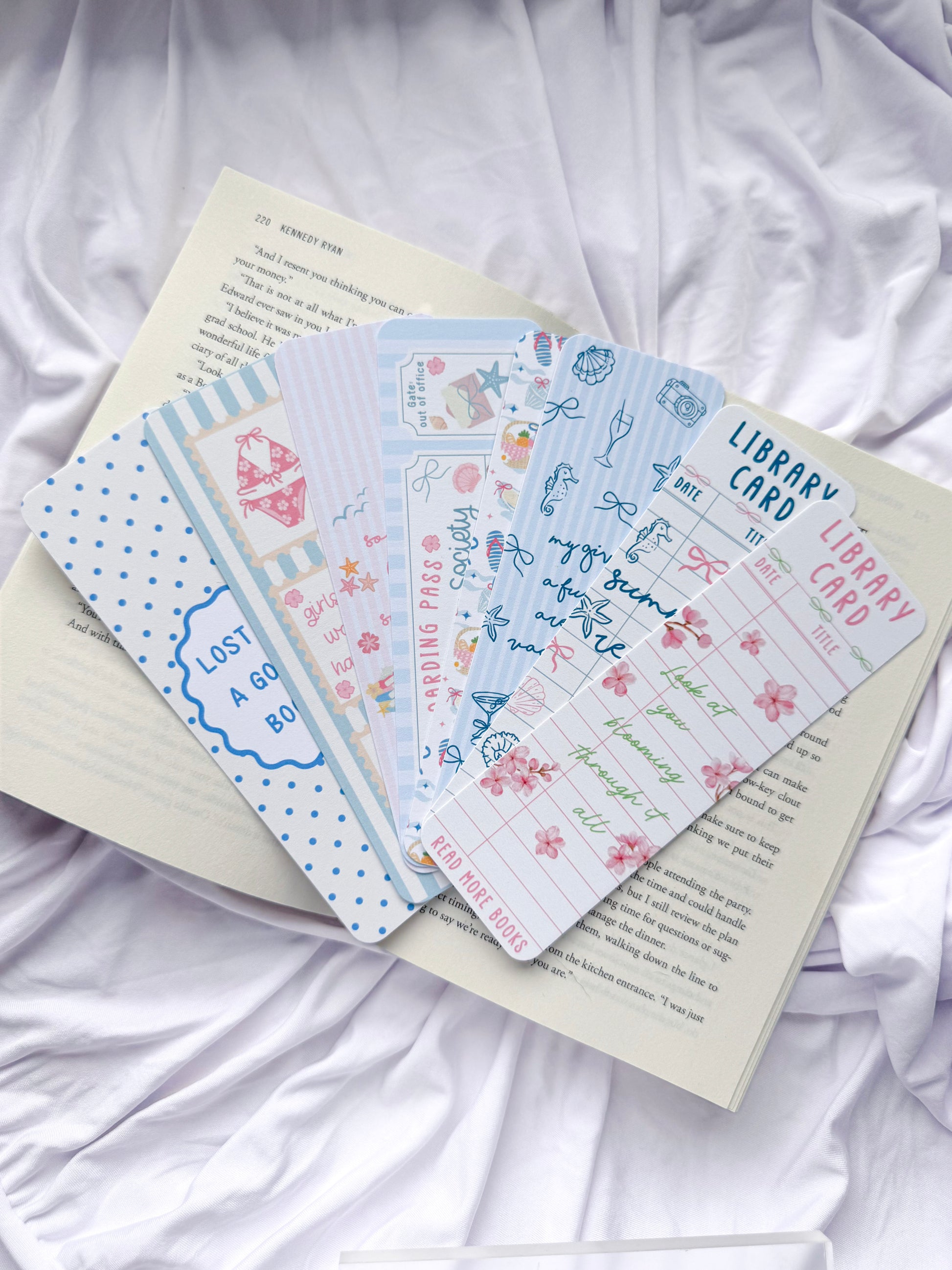 Flat lay of seven summer-themed bookmarks displayed on an open book with white bedding. The bookmark designs feature coastal illustrations, beach icons, pastel colors, library card designs, floral accents, and bookish quotes, creating the perfect reading accessories for summer book lovers.