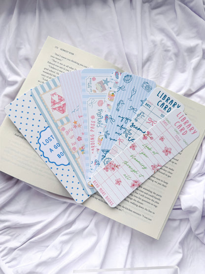 Flat lay of seven summer-themed bookmarks displayed on an open book with white bedding. The bookmark designs feature coastal illustrations, beach icons, pastel colors, library card designs, floral accents, and bookish quotes, creating the perfect reading accessories for summer book lovers.