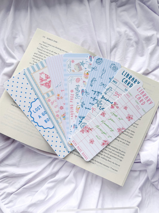 Flat lay of seven summer-themed bookmarks displayed on an open book with white bedding. The bookmark designs feature coastal illustrations, beach icons, pastel colors, library card designs, floral accents, and bookish quotes, creating the perfect reading accessories for summer book lovers.