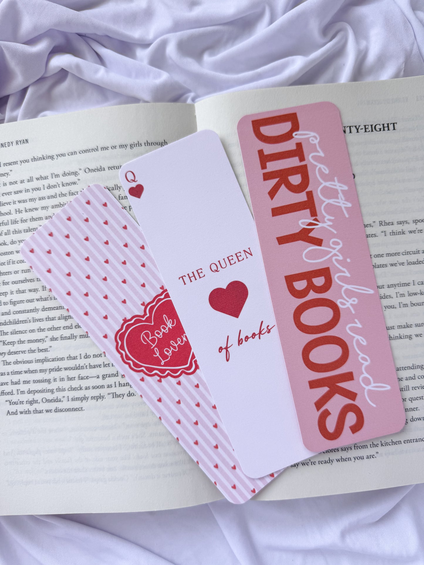 Flat lay of three pink romance-themed bookmarks displayed on an open book with white bedding. The bookmark designs feature a coquette heart pattern, "The Queen of Books" playing card design, and a "Dirty Girls Read Dirty Books" quote for romance readers
