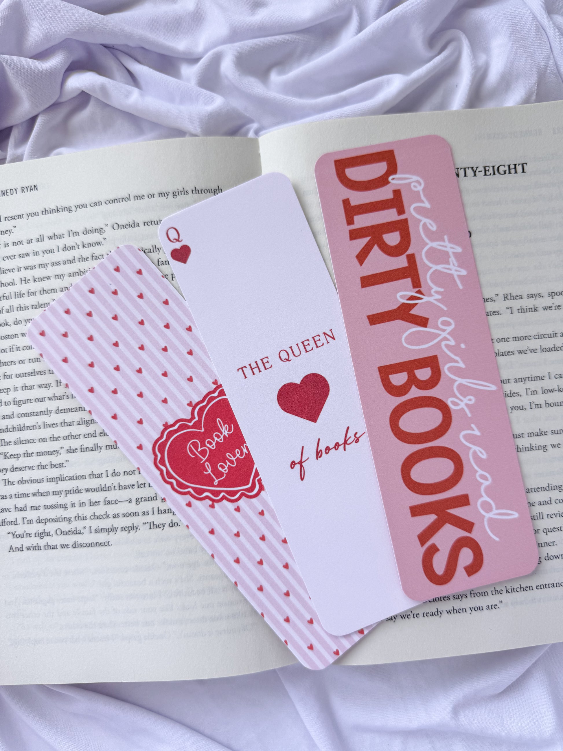 Flat lay of three pink romance-themed bookmarks displayed on an open book with white bedding. The bookmark designs feature a coquette heart pattern, "The Queen of Books" playing card design, and a "Dirty Girls Read Dirty Books" quote for romance readers