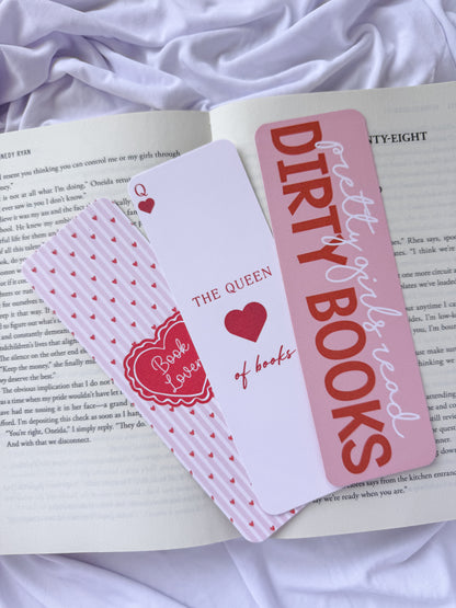 Flat lay of three pink romance-themed bookmarks displayed on an open book with white bedding. The bookmark designs feature a coquette heart pattern, "The Queen of Books" playing card design, and a "Dirty Girls Read Dirty Books" quote for romance readers