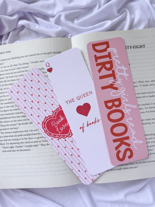 Flat lay of three pink romance-themed bookmarks displayed on an open book with white bedding. The bookmark designs feature a coquette heart pattern, "The Queen of Books" playing card design, and a "Dirty Girls Read Dirty Books" quote for romance readers